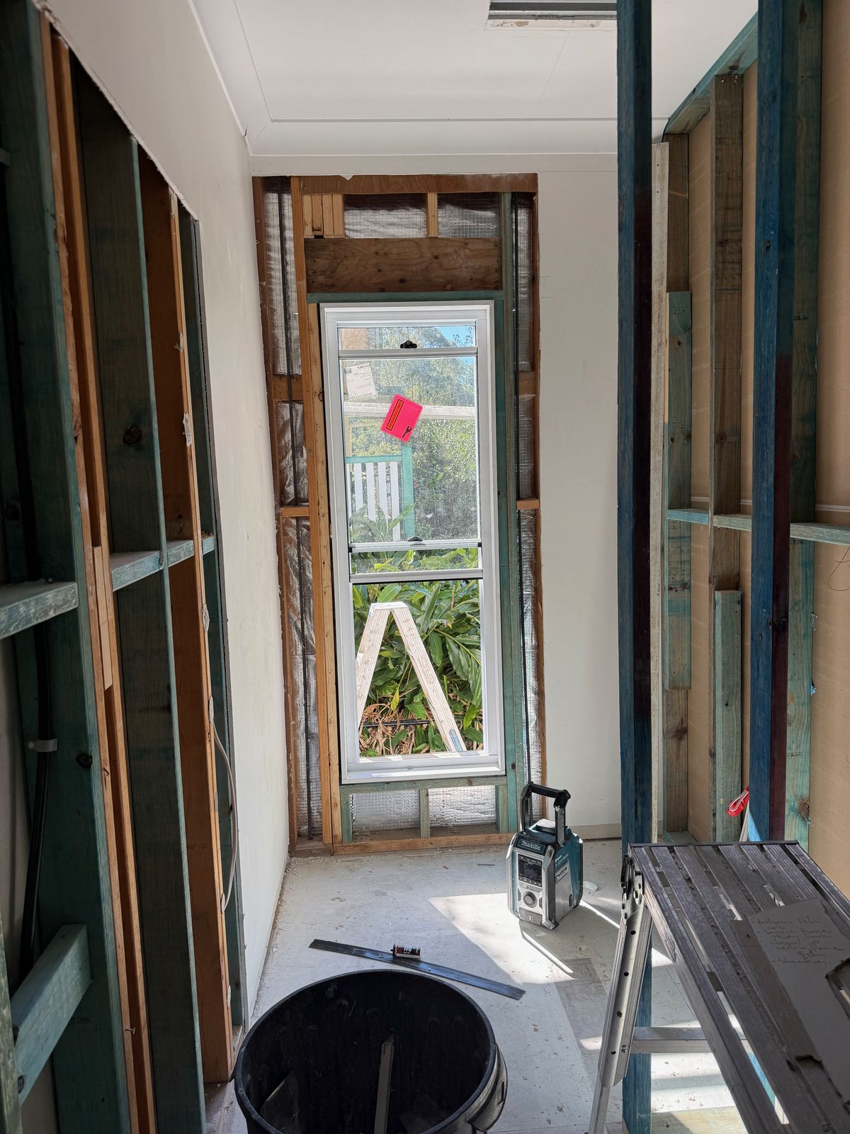 A bathroom in an old house stripped to the studs, the timber wall frame and back door opening exposed and a bucket on the bare floor