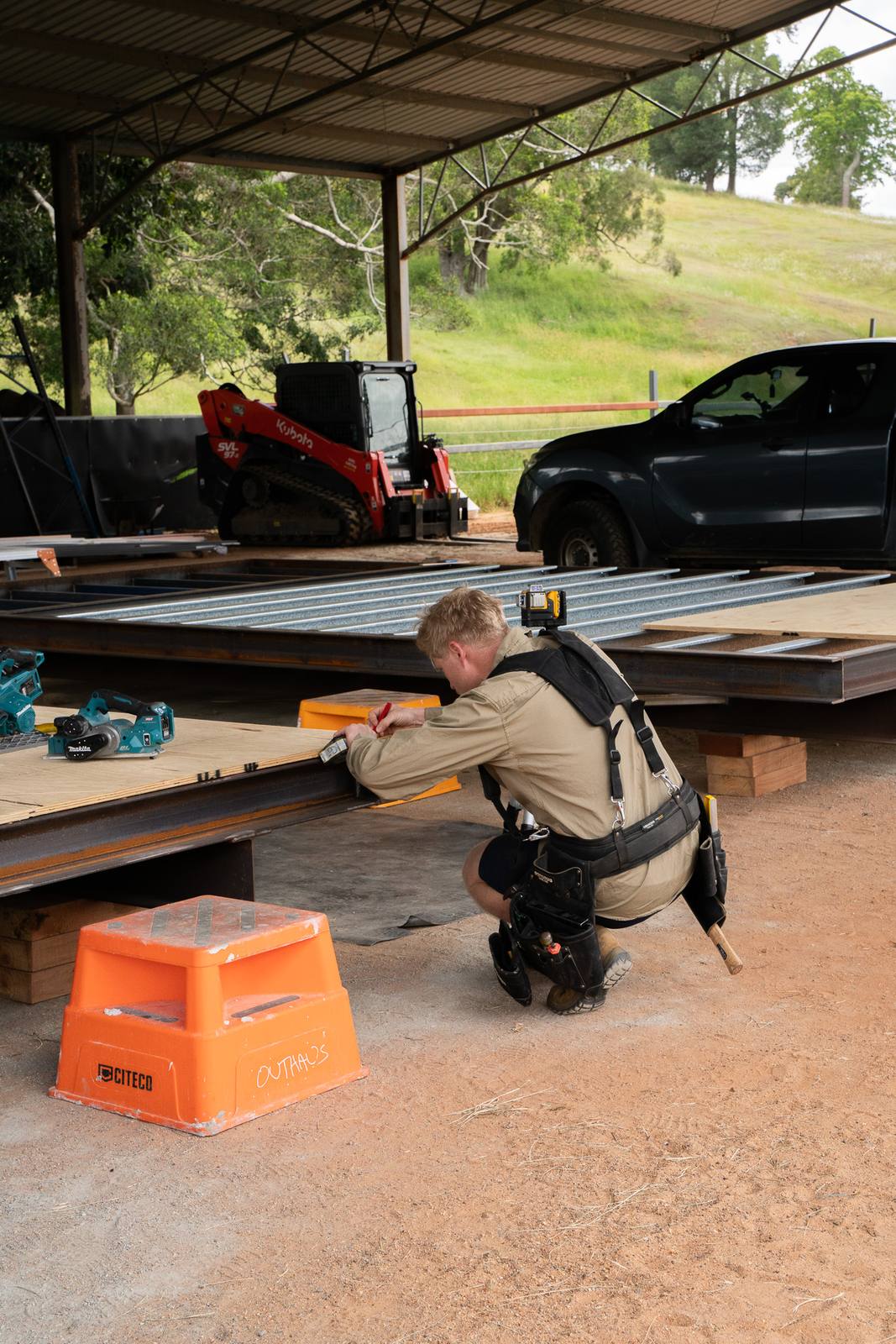 A carpenter in a tool belt kneeling on a module floor under the open shed, marking out the ply before it's fixed