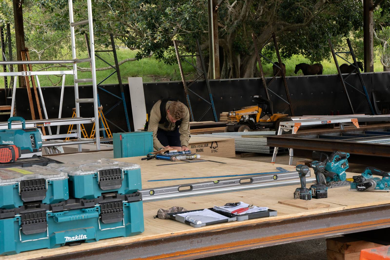 A builder measuring out on a ply floor panel, tool cases and a long level laid out on the bench