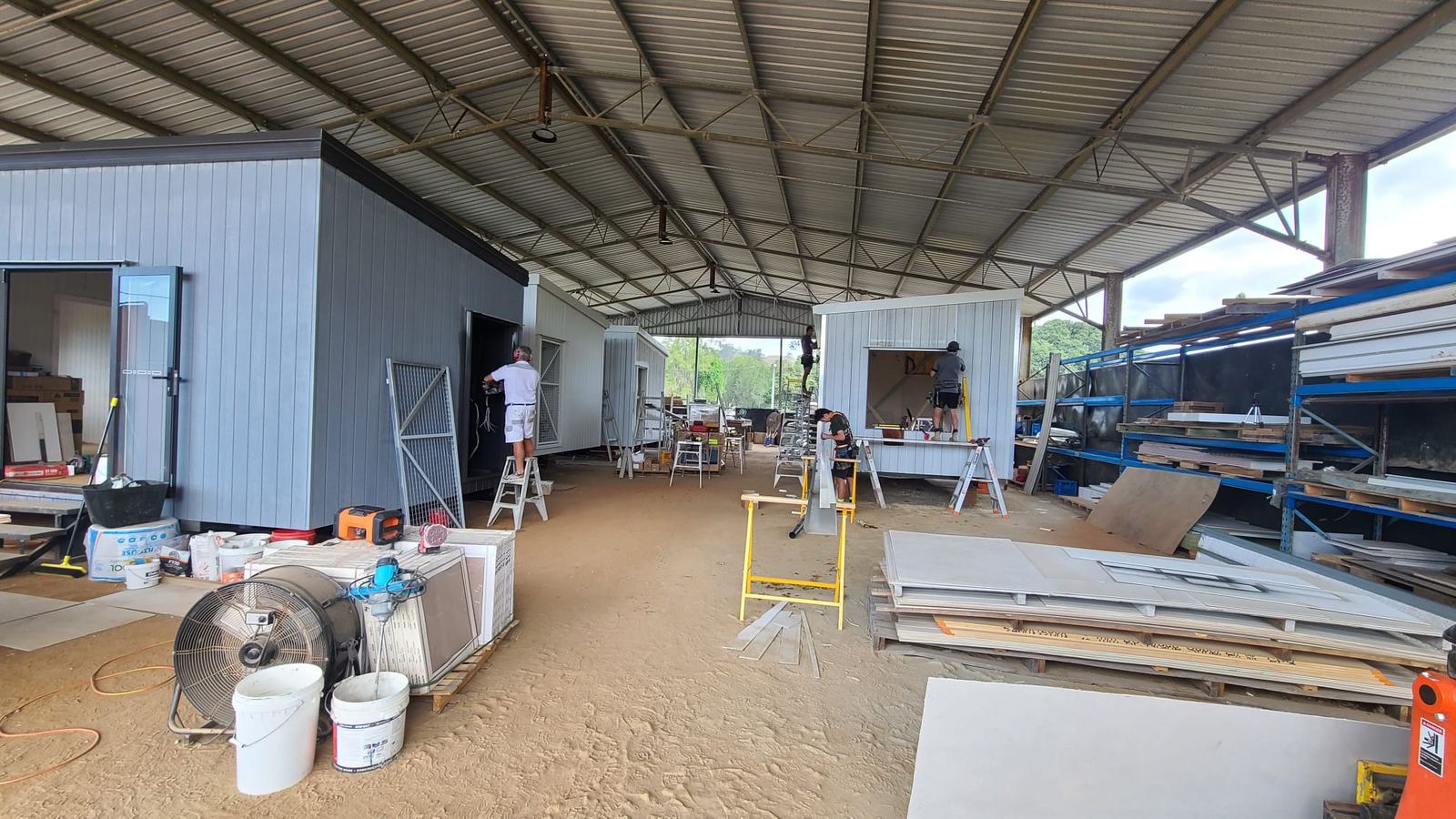 Looking down a long open shed with modules at different stages, sheet stock racked along one side and a builder at work
