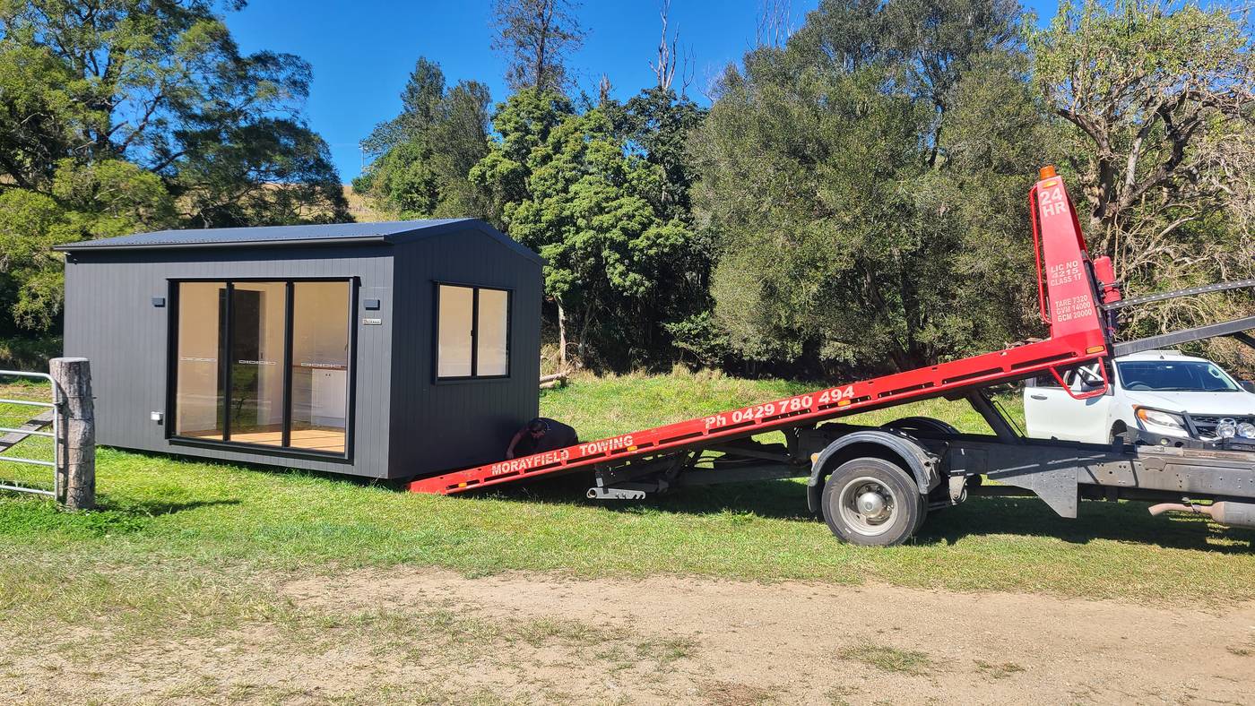 A dark-clad modular home on a red tilt tray truck at a farm gate, gum trees behind