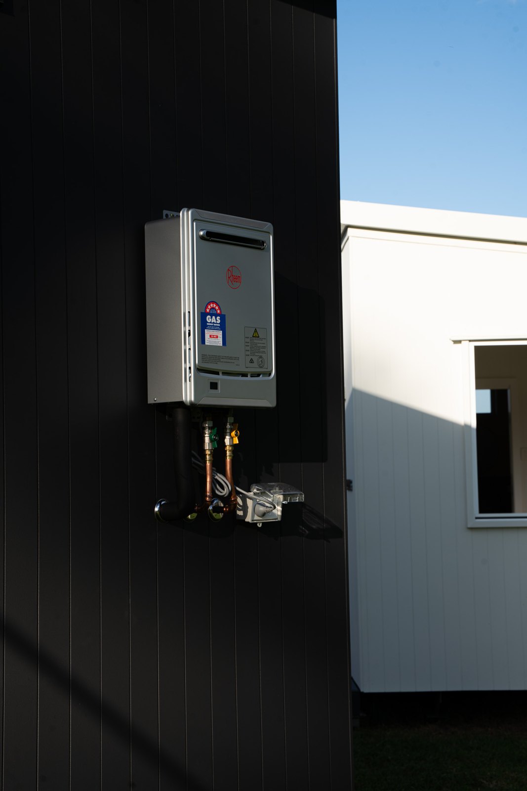 A gas instantaneous hot water unit mounted on the dark cladding of a second home, pipes and isolator below