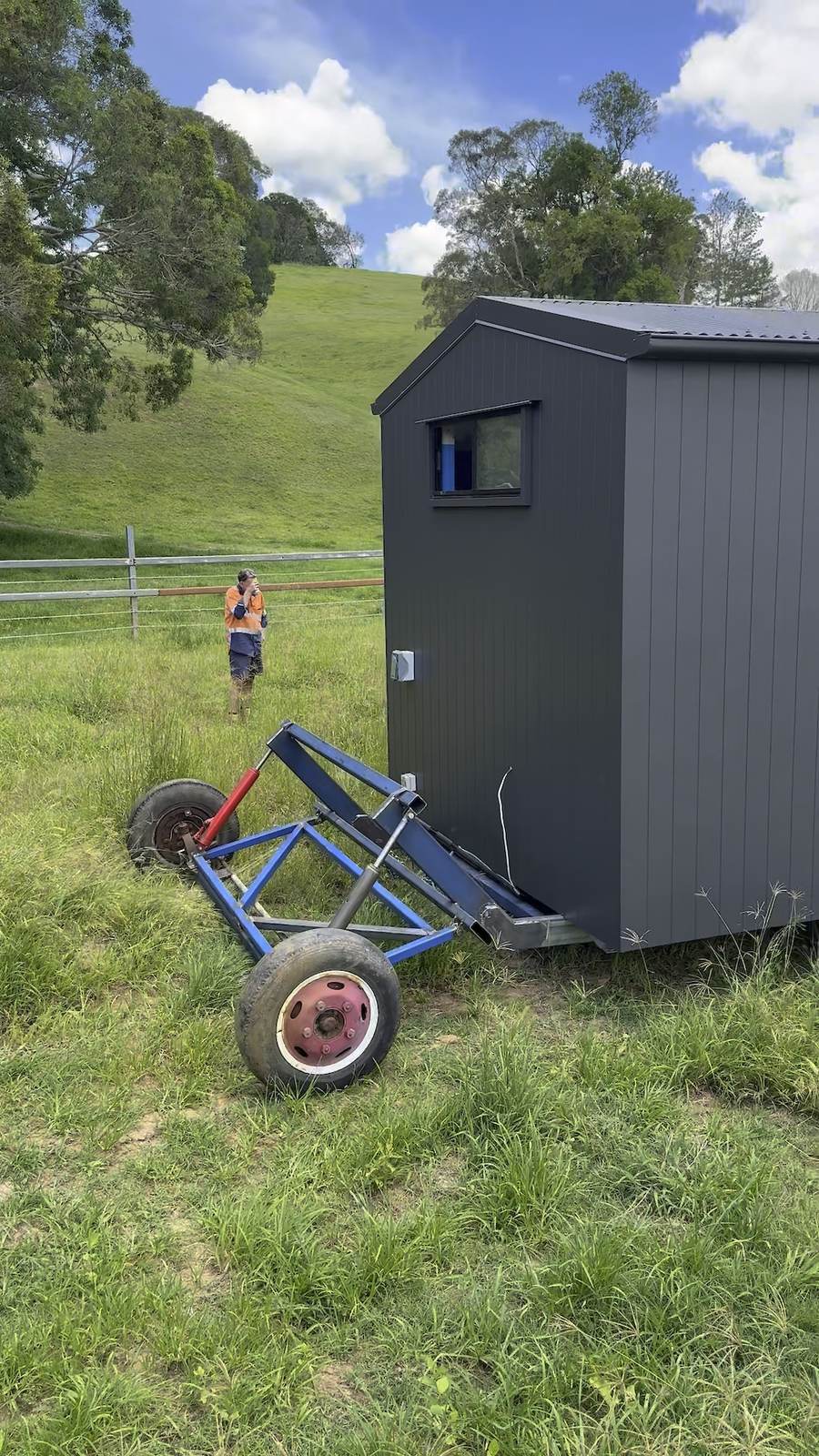 A dark-clad module on a steel transport dolly being moved across a paddock, a crew member watching from the fence