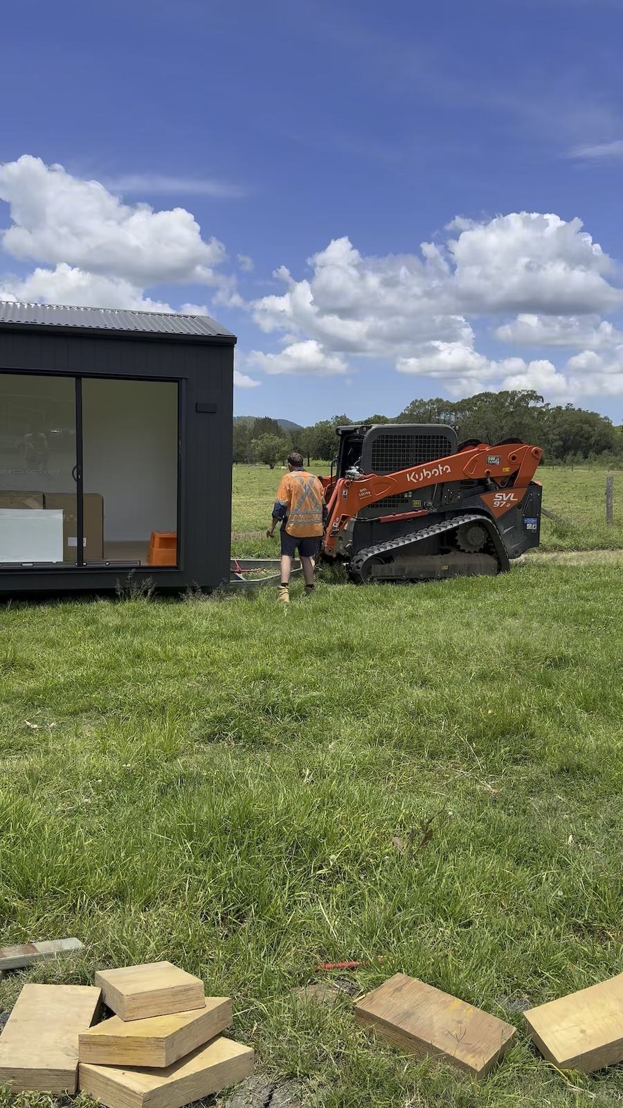 A dark-clad module set down on the grass beside a skid steer, timber packers stacked in the foreground