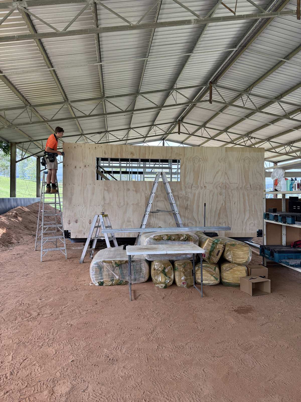 A builder on a ladder fixing ply sheathing to a module in the shed, insulation bags stacked in front
