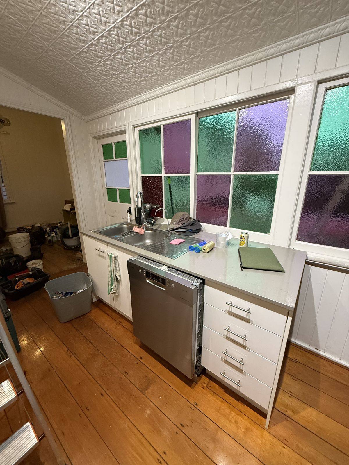 The old kitchen in a Queenslander, white cabinets under coloured leadlight windows and a pressed metal ceiling