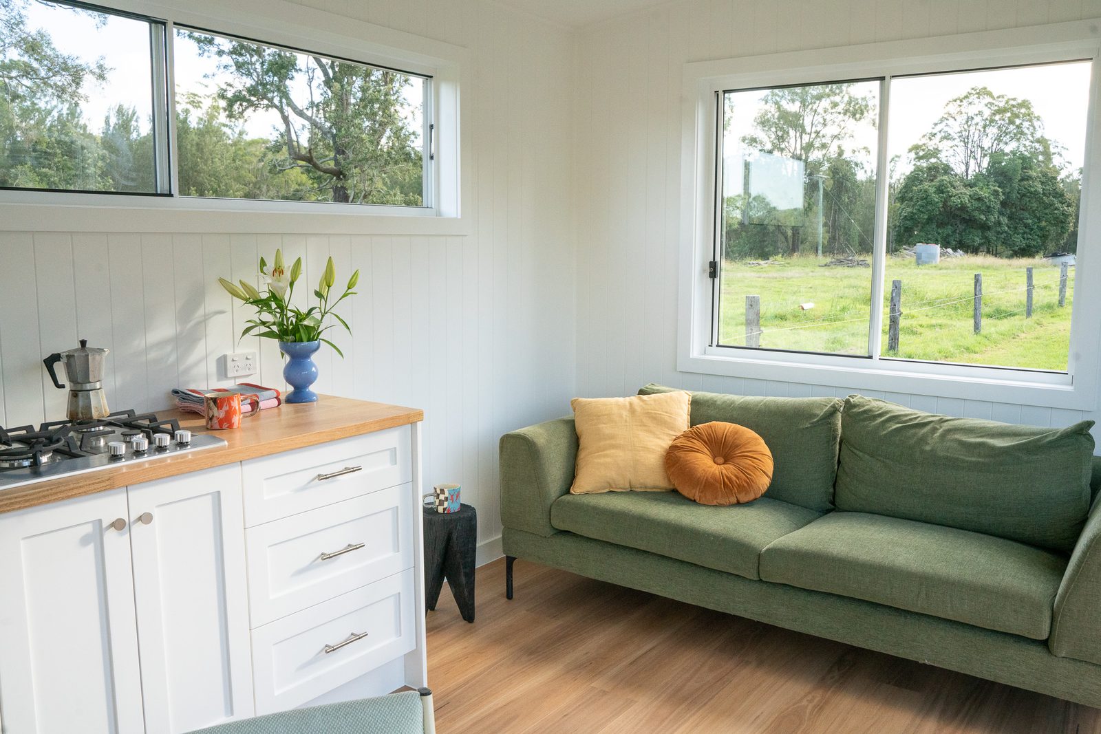 Inside a white second home: kitchen with timber benchtop and a green sofa under two windows
