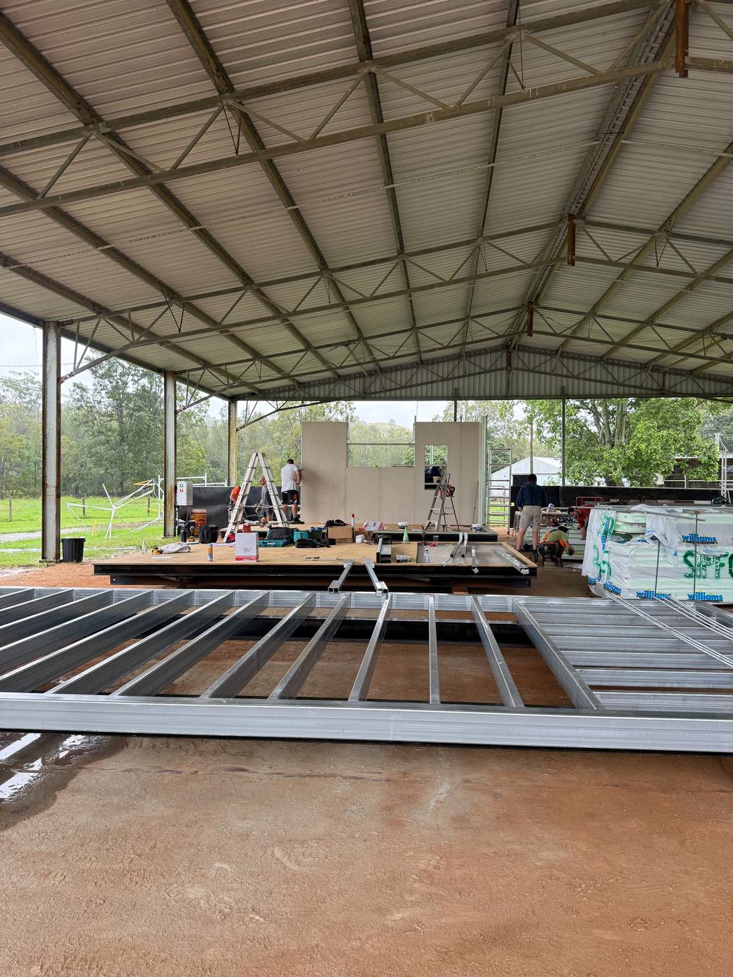 Galvanised steel floor frames laid out on the dirt under a big open shed, a module being fitted out behind