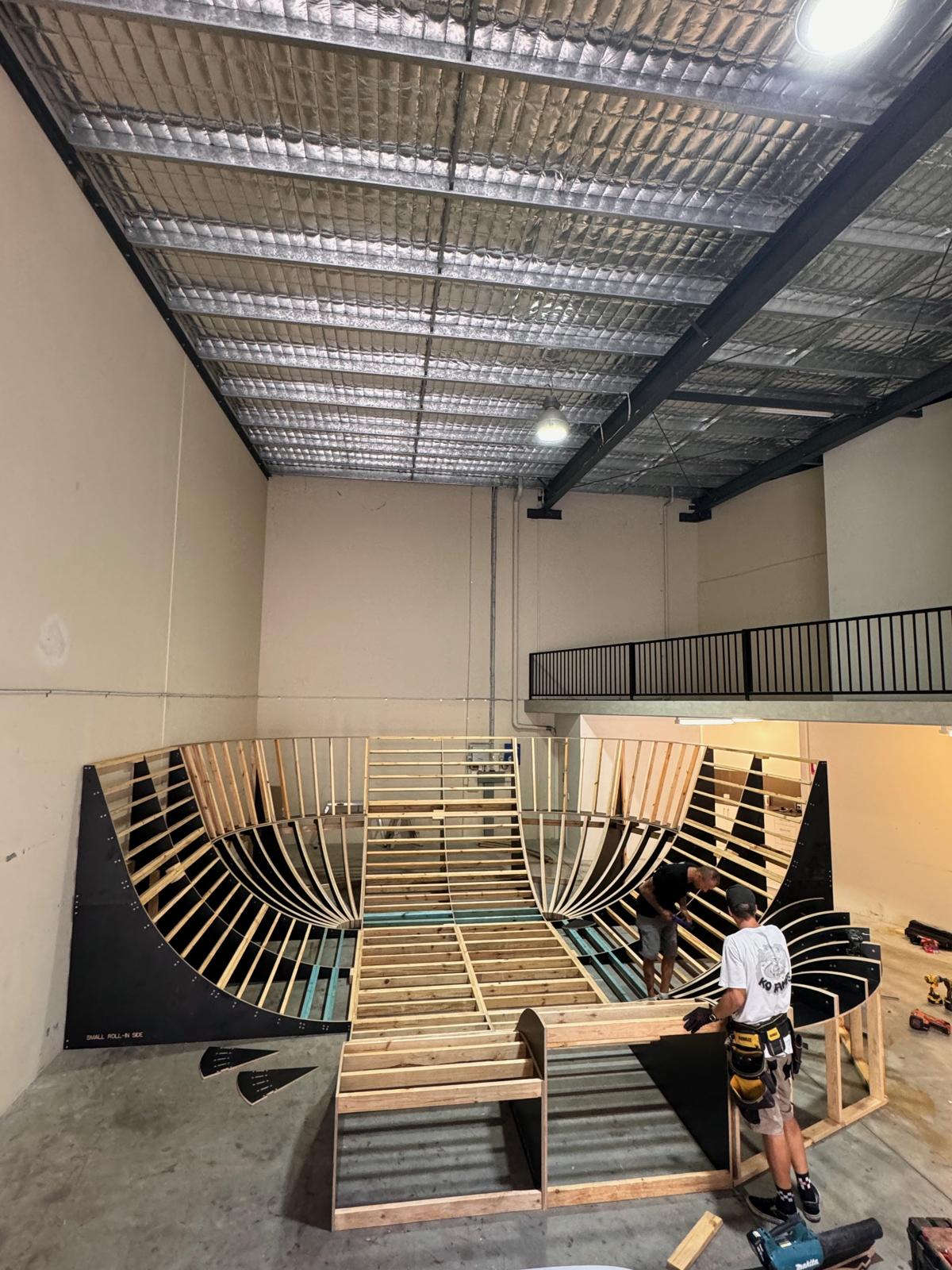 A carpenter building the timber frame of a large indoor skate ramp in a warehouse