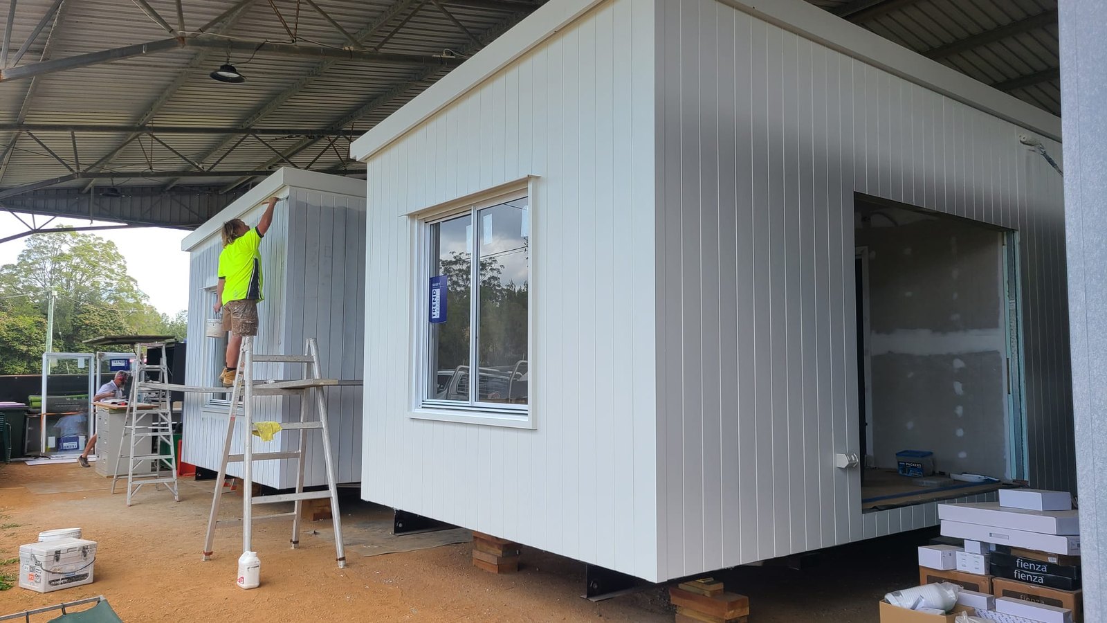 Two white-clad modules on stands in the shed, a painter in hi-vis on a ladder finishing the cladding